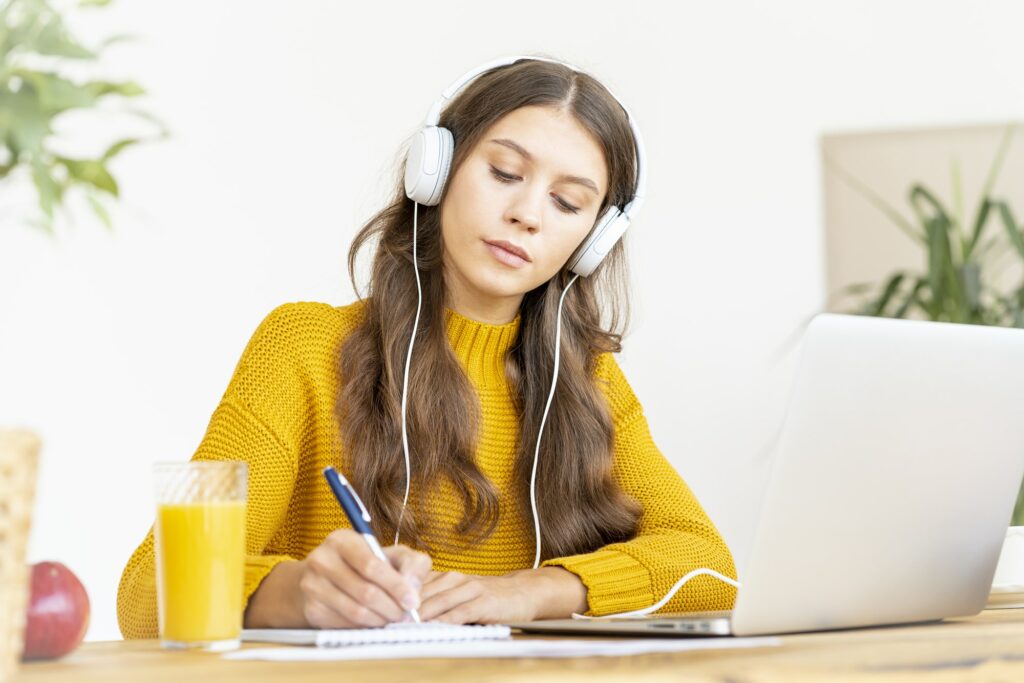 Happy female in wireless headphones studying online course, using pc and writing in notepad